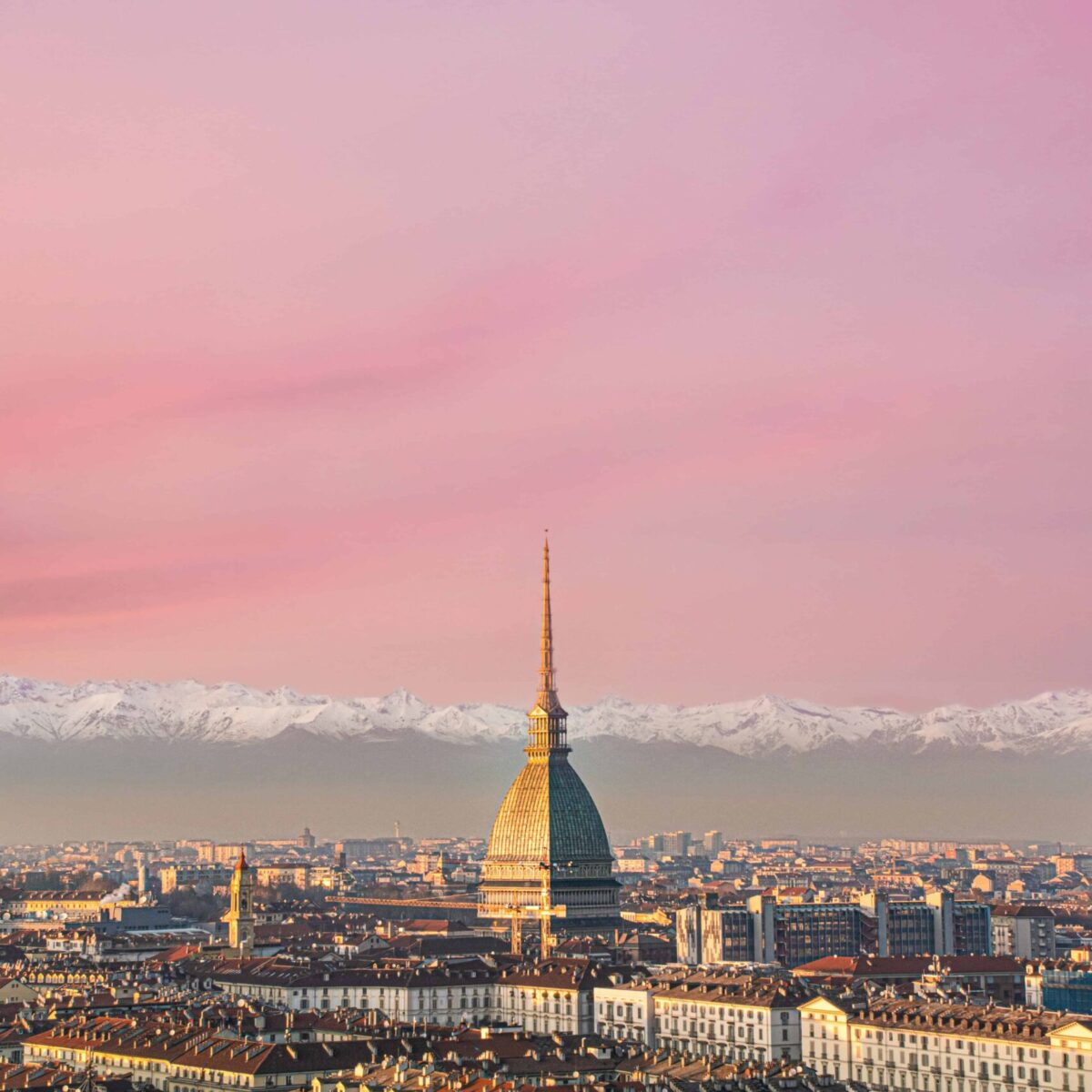 Widok na Turyn z Doliny Południowej, z charakterystyczną kopułą Mole Antonelliana i górami Alp w tle, podczas zachodu słońca, malujący miasto w ciepłych odcieniach różu.