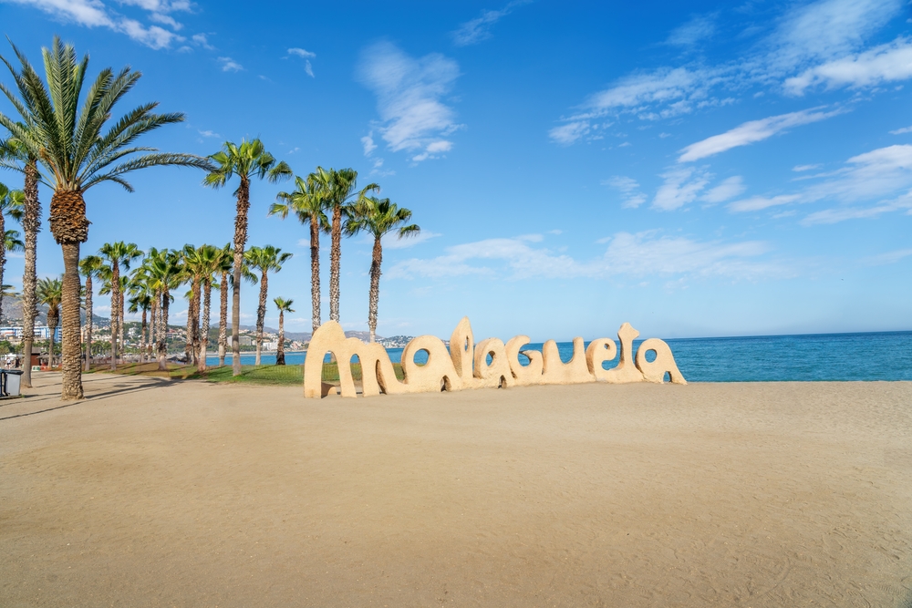 Piaskowa rzeźba z napisem Malagueta na plaży w Maladze, za nią rząd palm i morze