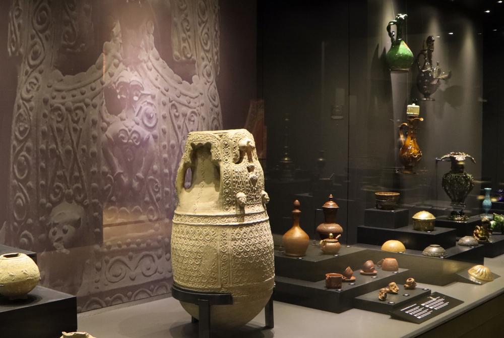 Ekspozycja muzealna z ozdobnymi ceramicznymi naczyniami i dzbanami oraz rzeźbionym kamiennym reliefem w tle w Akarze