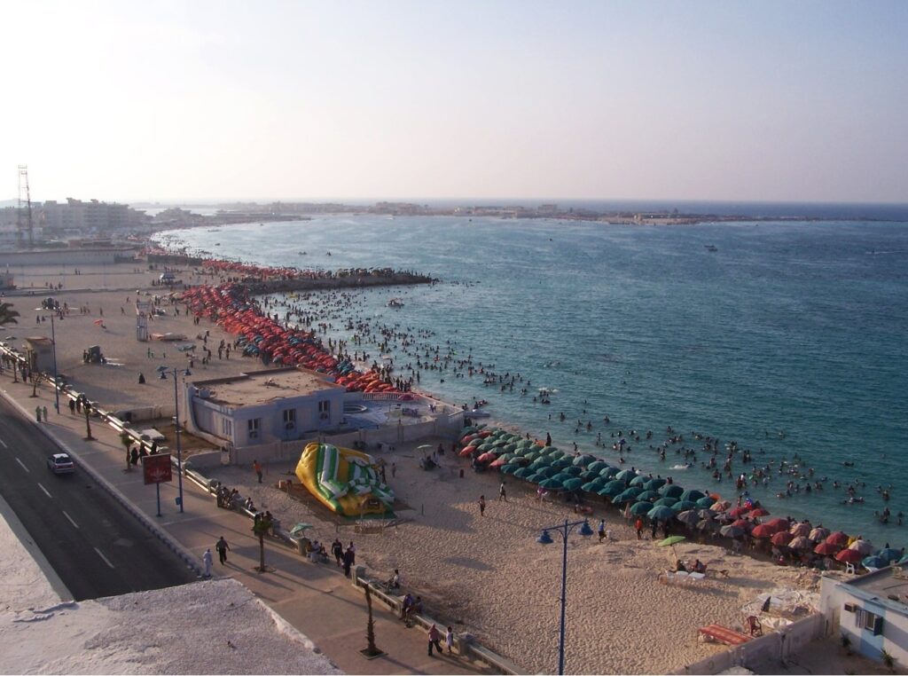 Miejska plaża w Marsa Matruh w szczycie sezonu, rzędy parasoli i kąpiący się w płytkiej zatoce