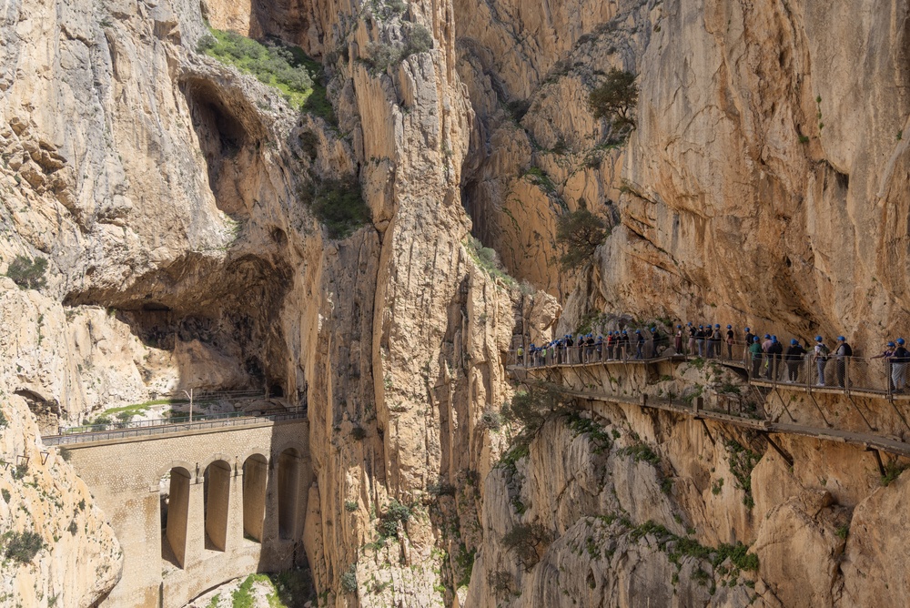 Kładka Caminito del Rey na ścianie wąwozu, w dole kamienny wiadukt kolejowy z arkadami