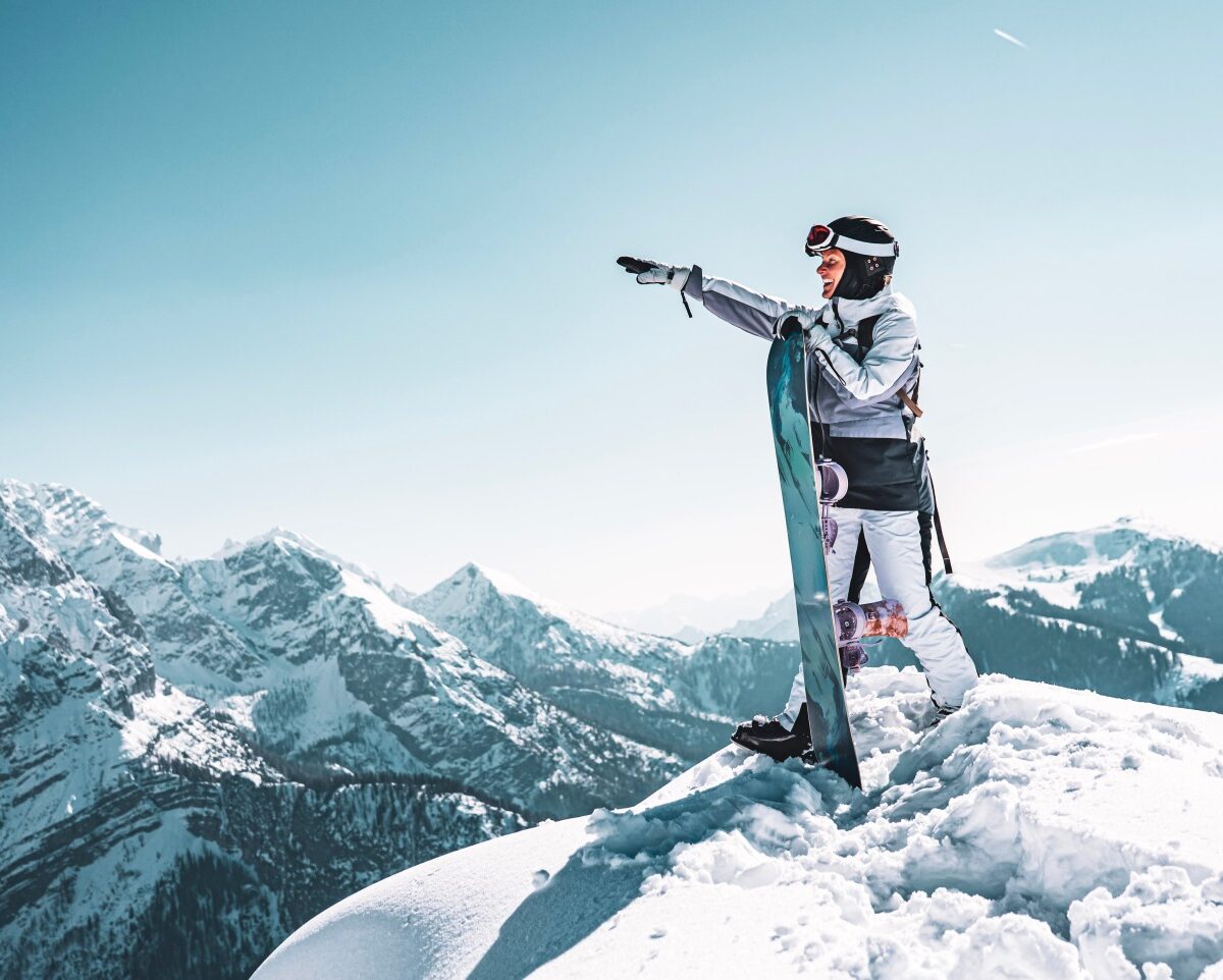 Snowboardzistka na ośnieżonym szczycie wskazuje ręką pasmo gór w słońcu