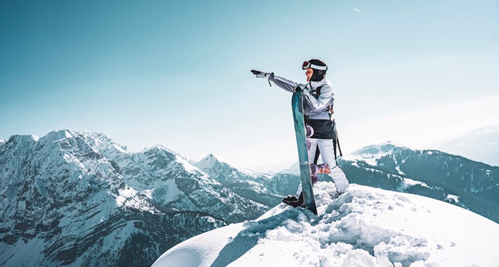 Snowboardzistka z deską na szczycie wskazuje ręką ośnieżone pasmo gór