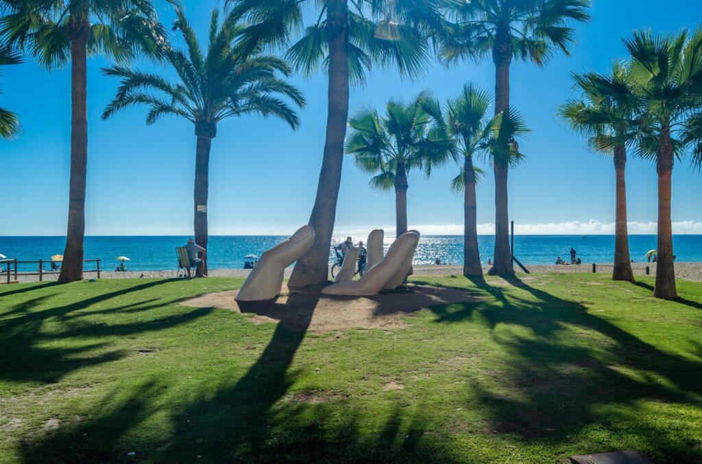 Rzeźba dłoni na trawniku przy plaży w Fuengiroli, rząd palm i morze w tle