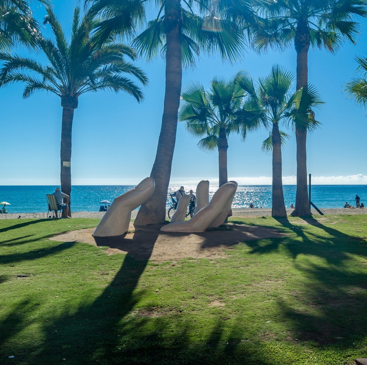 Rzeźba dłoni na trawniku przy plaży w Fuengiroli, rząd palm i morze w tle