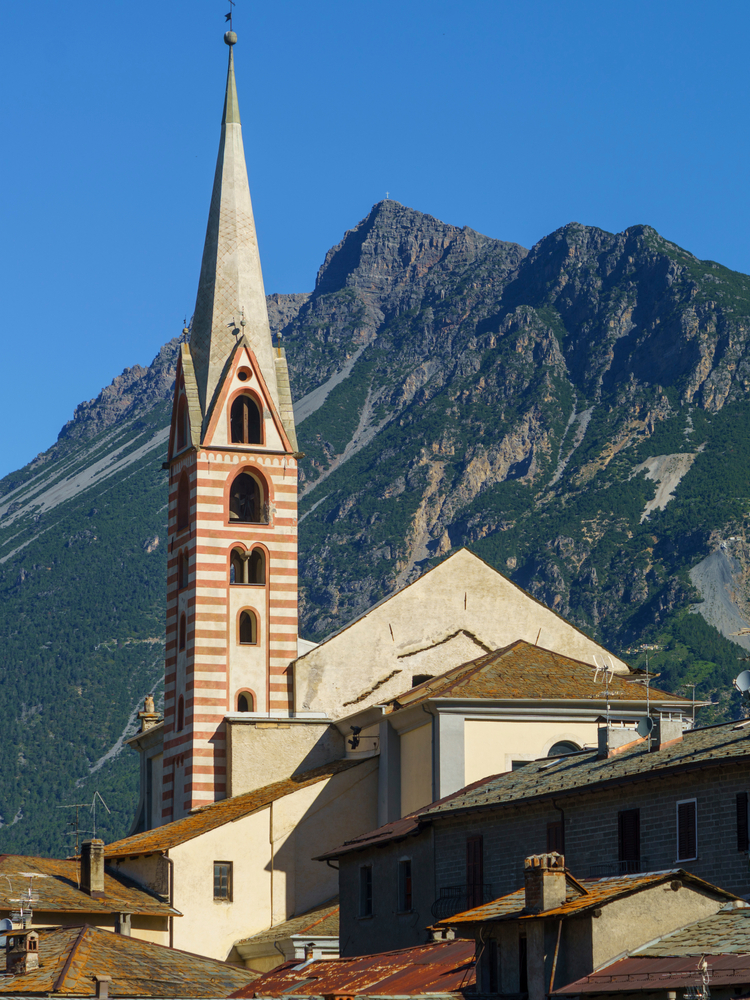 Pasiasta wieża kościoła nad dachami Bormio, w tle skaliste zbocze