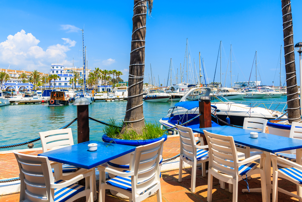 Stoliki nadbrzeżnej restauracji w porcie Estepony, jachty przy kei i palmy nad tarasem