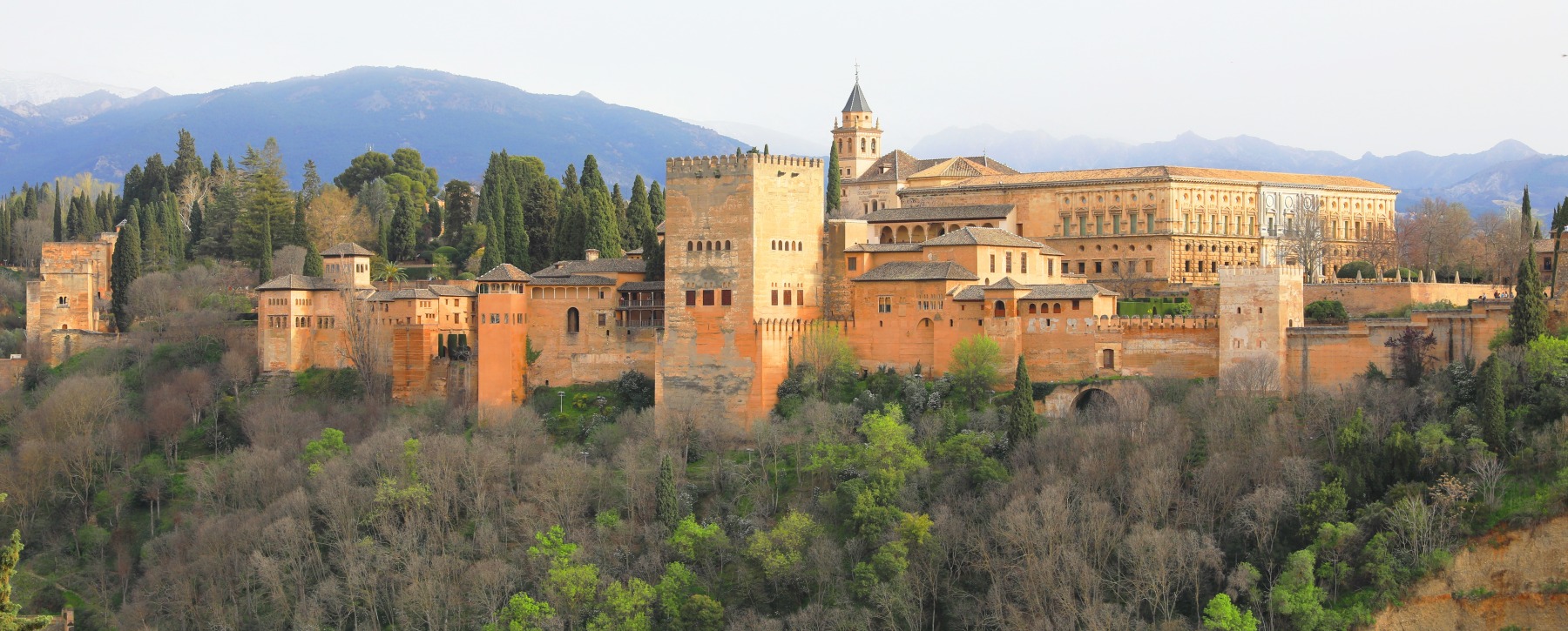 Alhambra w Grenadzie na zalesionym wzgórzu, ochrowe mury i wieża Comares, po prawej renesansowy pałac Karola V, w tle pasmo górskie