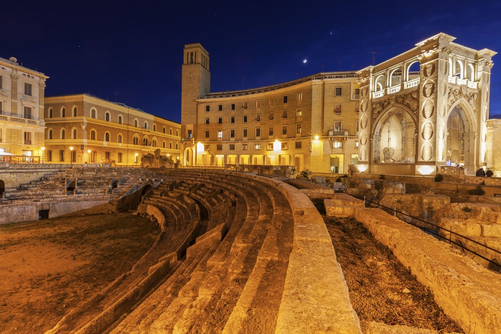 Rzymski amfiteatr poniżej poziomu Piazza Sant'Oronzo w Lecce nocą, nad nim podświetlone budynki placu