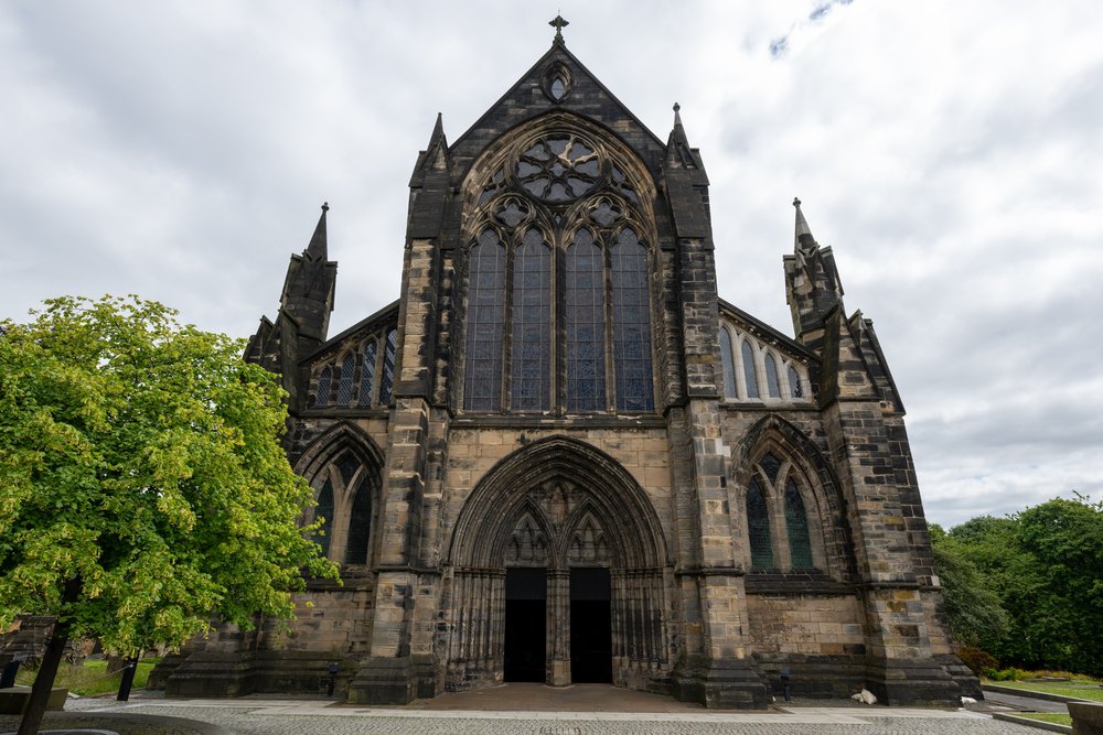 Gotycka fasada katedry w Glasgow z wysokim oknem nad wejściem, obok zielone drzewo