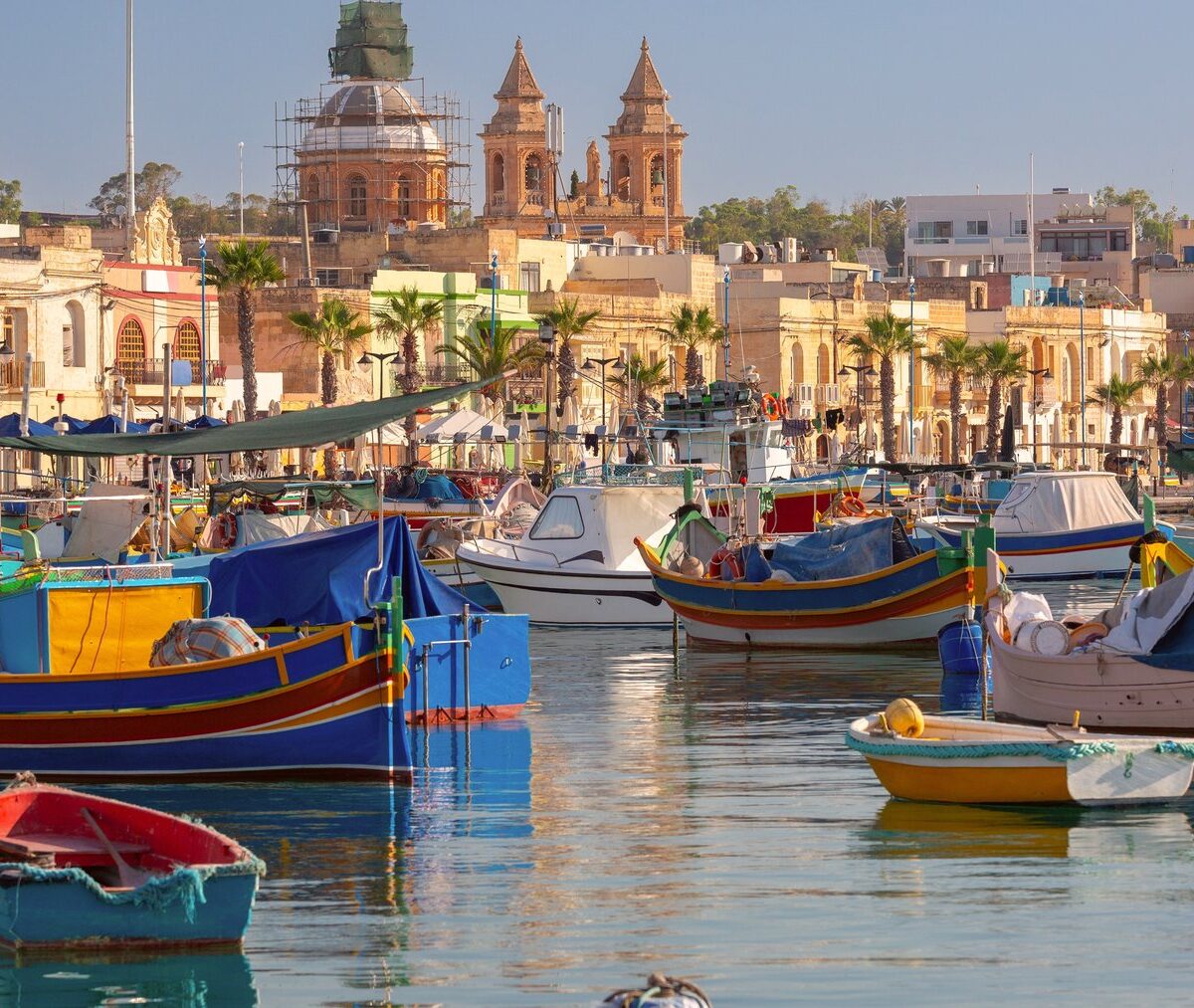 Kolorowe łodzie luzzu w porcie Marsaxlokk, w tle wieże kościoła parafialnego i palmy
