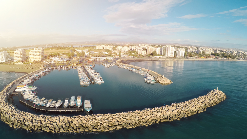 Marina w Larnace z jachtami za kamiennym falochronem, w tle plaża Finikoudes i miasto