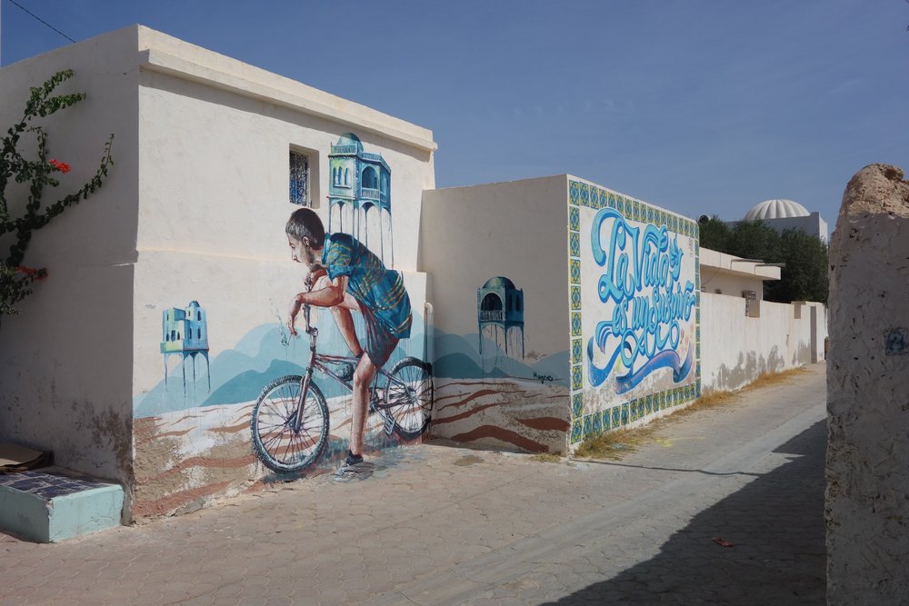 Mural chłopca na rowerze na ścianie domu w Erriadh, jedna z prac projektu Djerbahood, obok ściana z kaligrafią