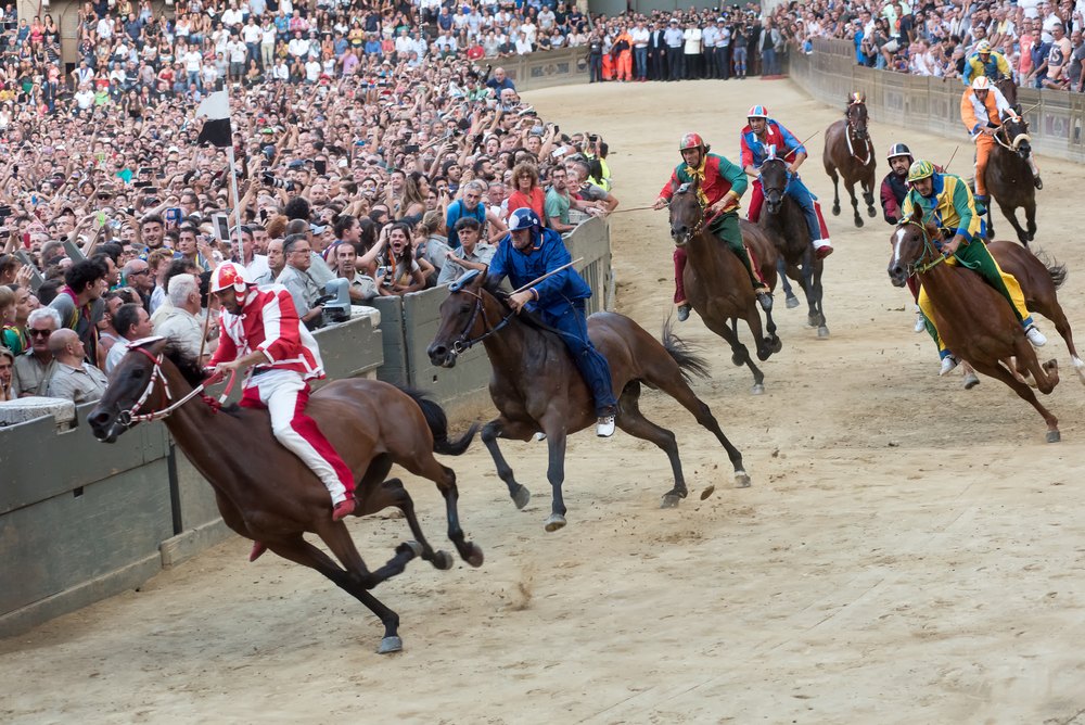 Dżokeje na koniach w barwach kontrad pędzą po torze na Piazza del Campo podczas Palio, za barierami tłum widzów