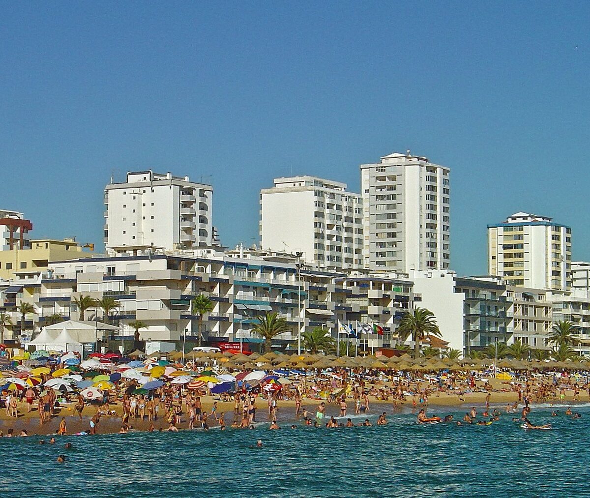 Plaża miejska Quarteiry z kolorowymi parasolami, za nią bloki i palmy wzdłuż promenady
