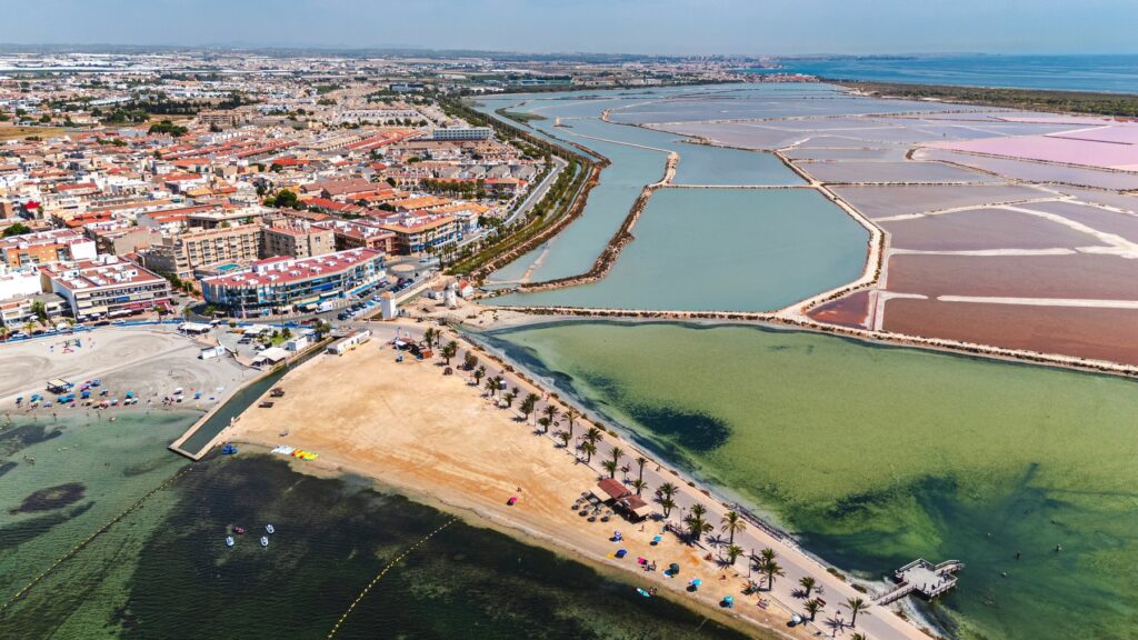 Lo Pagán nad Mar Menor z lotu ptaka: plaża z palmami na grobli, płytka laguna i różowe saliny San Pedro del Pinatar