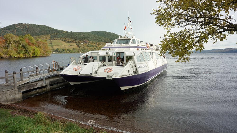Biały statek wycieczkowy przy pomoście nad Loch Ness pod zalesionym wzgórzem