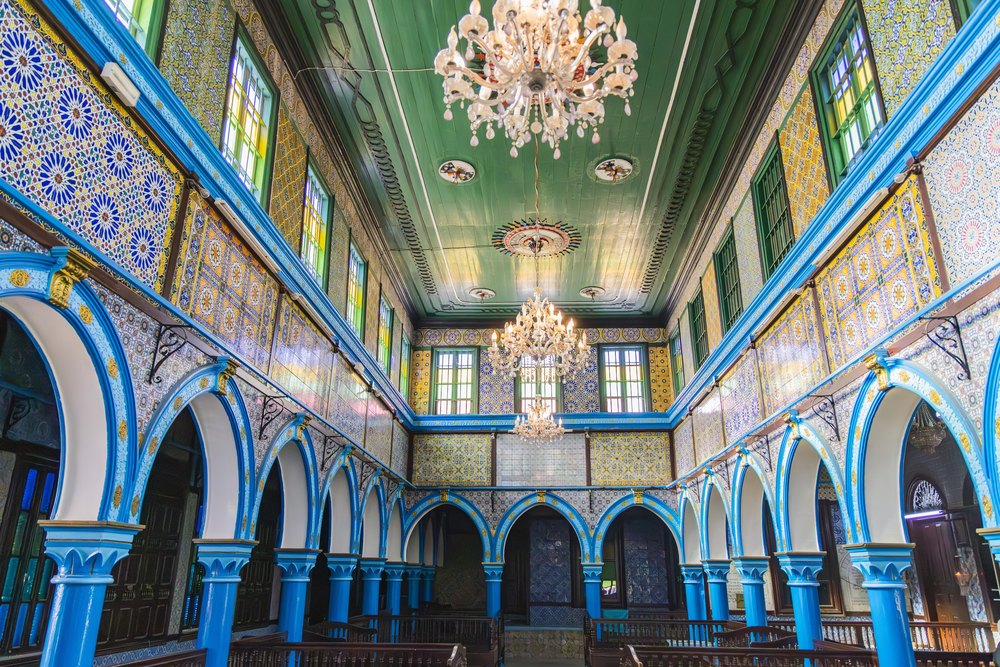 Wnętrze synagogi El Ghriba na Dżerbie: niebieskie łuki na kolumnach, ściany z kafli i kryształowe żyrandole