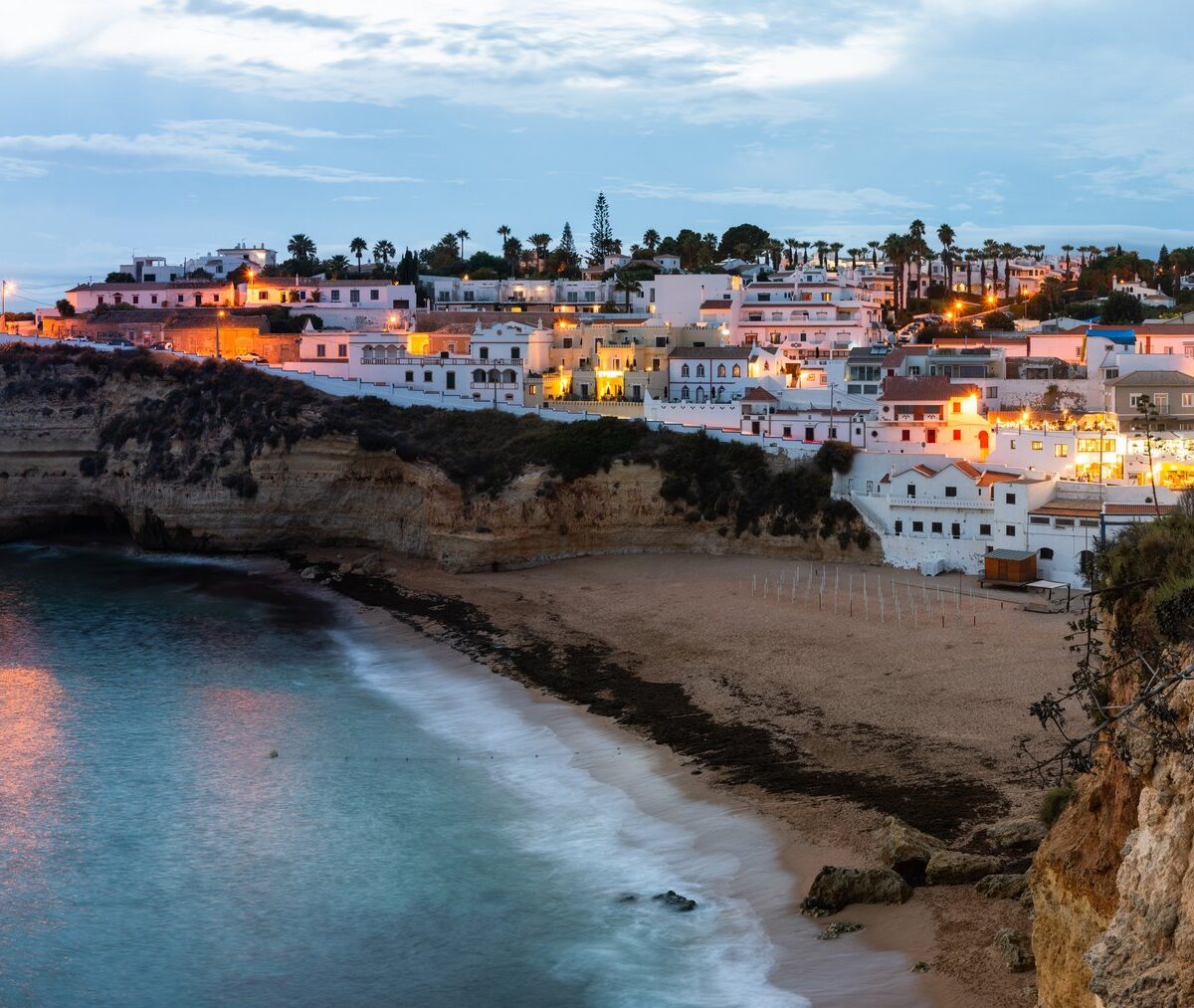 Białe domy Carvoeiro na klifie nad zatoką z plażą, o zmierzchu z zapalonymi światłami